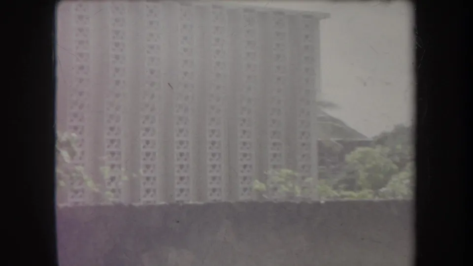 1969 Hawaii home-movie frame showing a tall building beyond trees and a wall, with soft focus and dark 8mm frame borders.