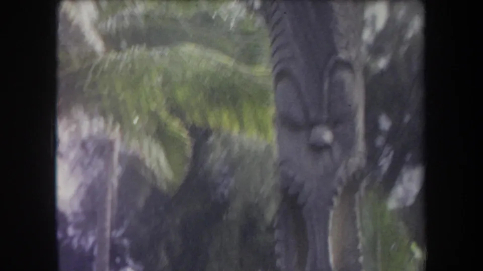 1969 Hawaii home-movie view of a large tiki statue partly framed by palms and dense tropical foliage.