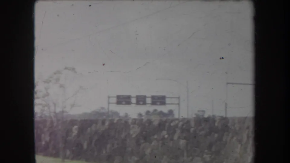 1969 Hawaii 8mm frame with a distant roadside sign, stone wall, bare branches, and an overcast sky.