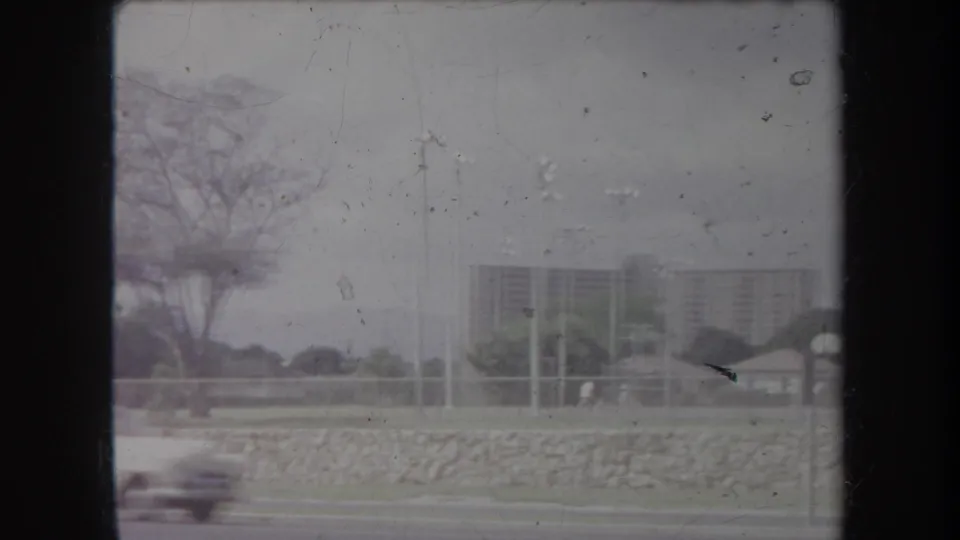 1969 Hawaii drive-by frame showing a blurred car, roadside stone wall, tall light poles, trees, and distant high-rise buildings.