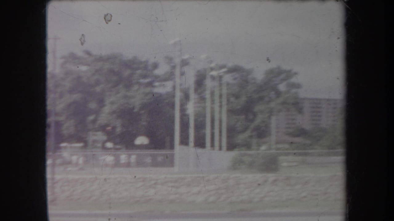 1969 Hawaii home-movie frame showing flagpoles, trees, low walls, and distant buildings in soft faded color.