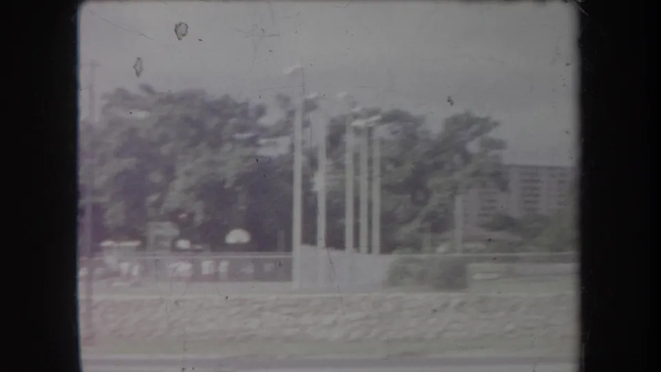 1969 Hawaii home-movie frame showing flagpoles, trees, low walls, and distant buildings in soft faded color.