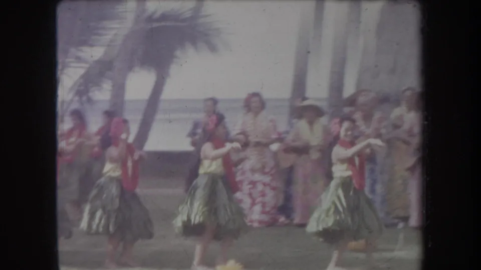 1969 Hawaii home-movie frame showing hula dancers with green skirts and red flower accessories performing near palm trees and spectators.
