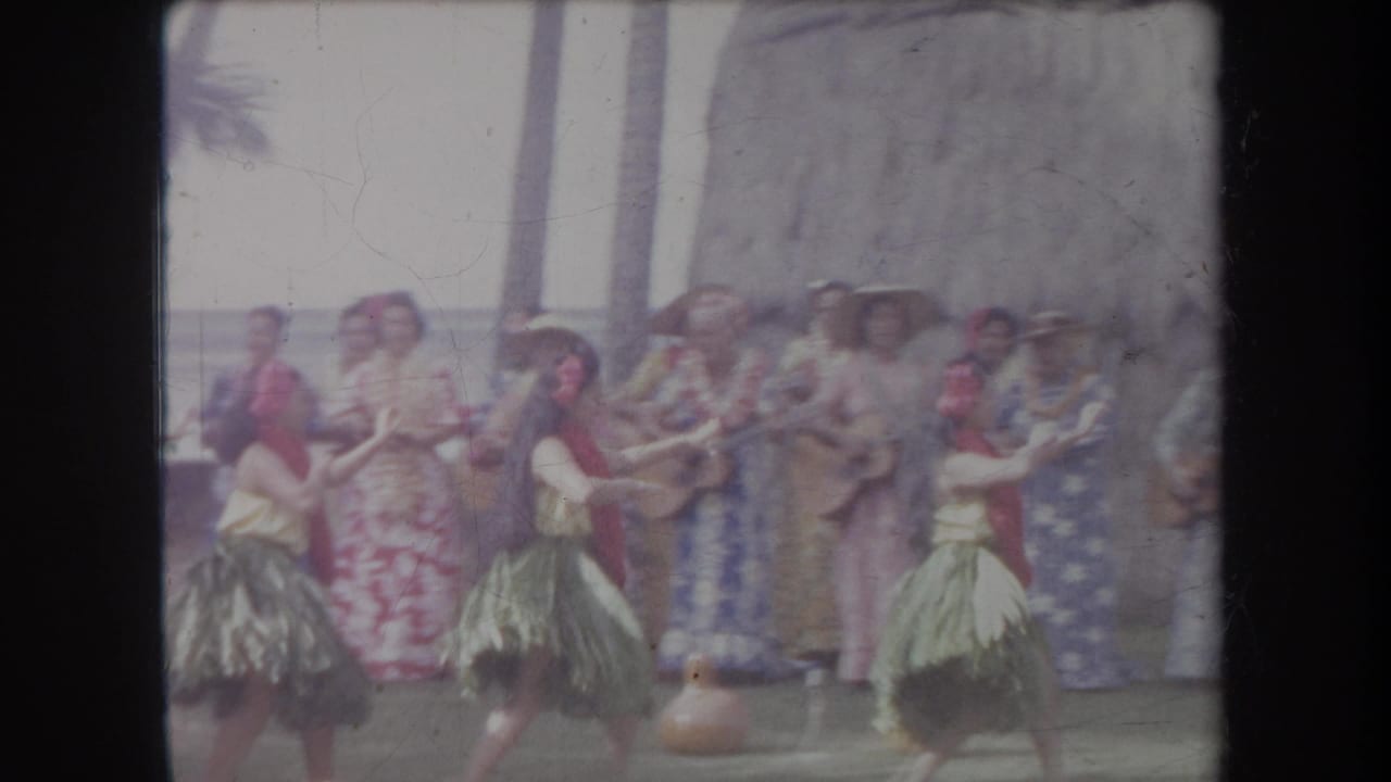 1969 Hawaii home-movie frame showing hula dancers in grass skirts and red leis performing before tourists near a thatched hut.