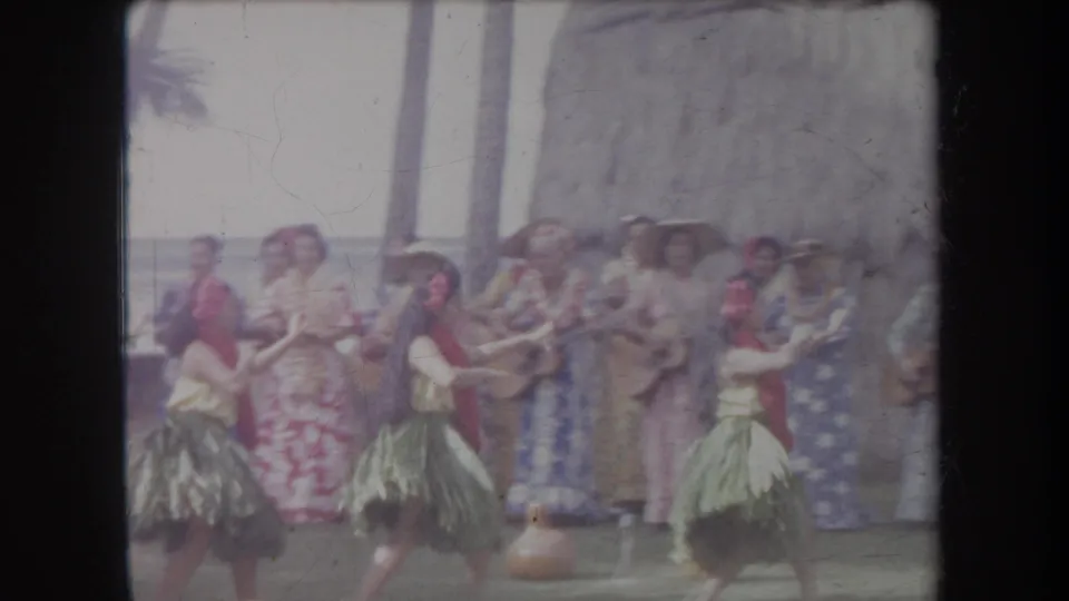 1969 Hawaii home-movie frame showing hula dancers in grass skirts and red leis performing before tourists near a thatched hut.