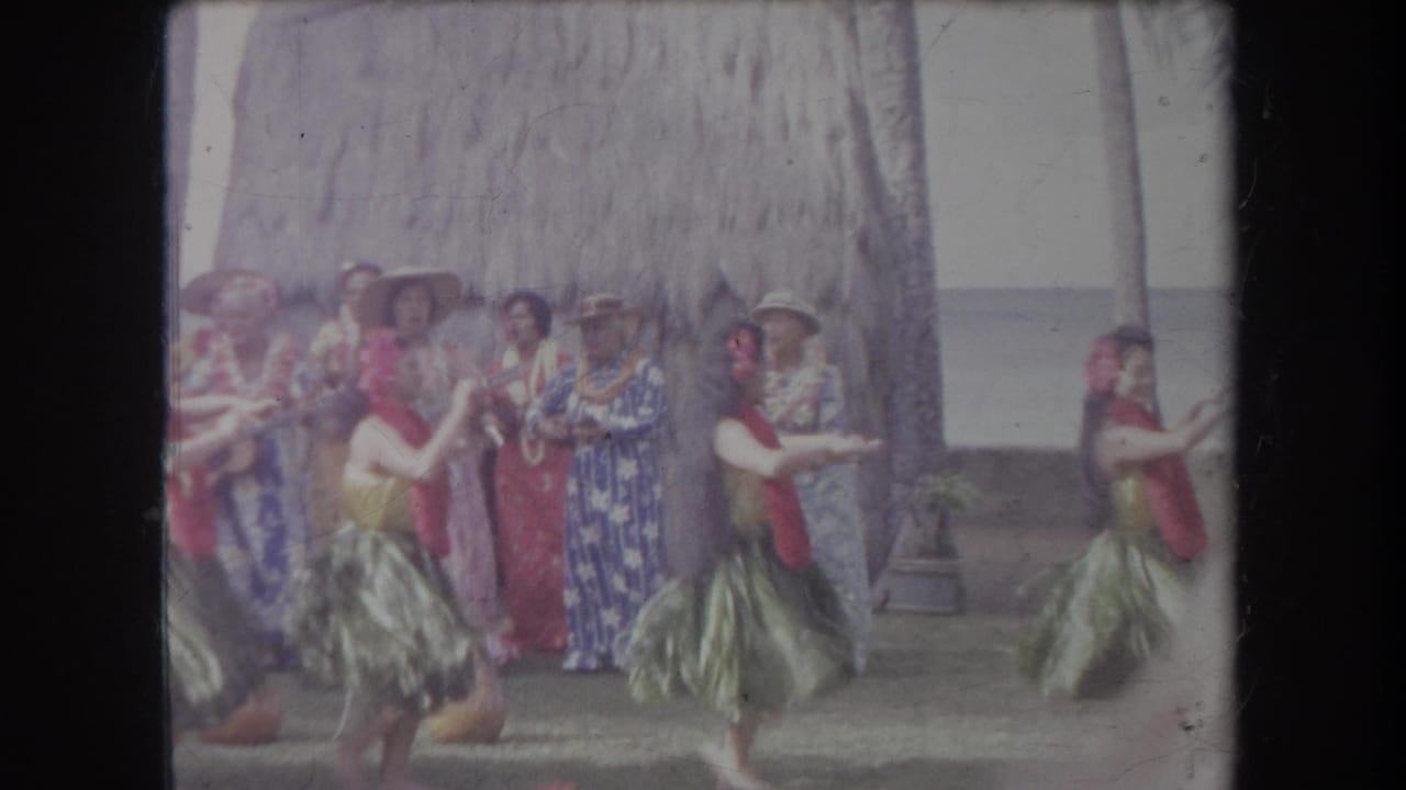 Dancers in grass skirts pass in front of a group of spectators by a thatched hut.