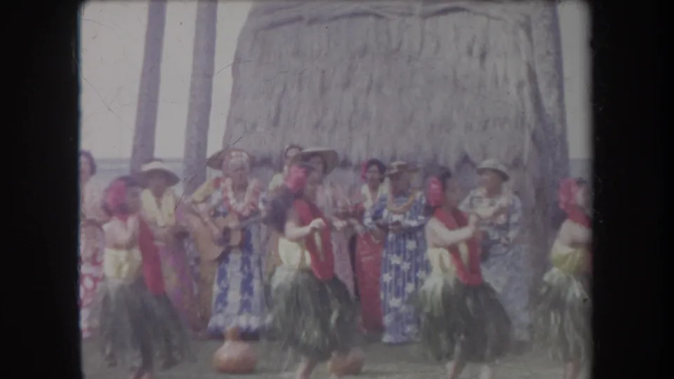 Soft 8mm Hawaii vacation frame showing hula dancers and tourists grouped near a thatched hut during a beach performance.