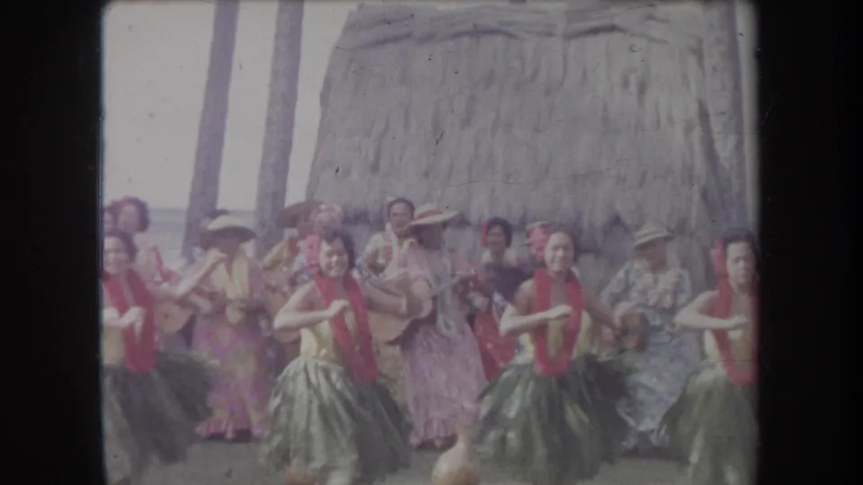 1969 Hawaii hula performance frame with dancers facing forward, tourists behind them, and a thatched hut in the background.