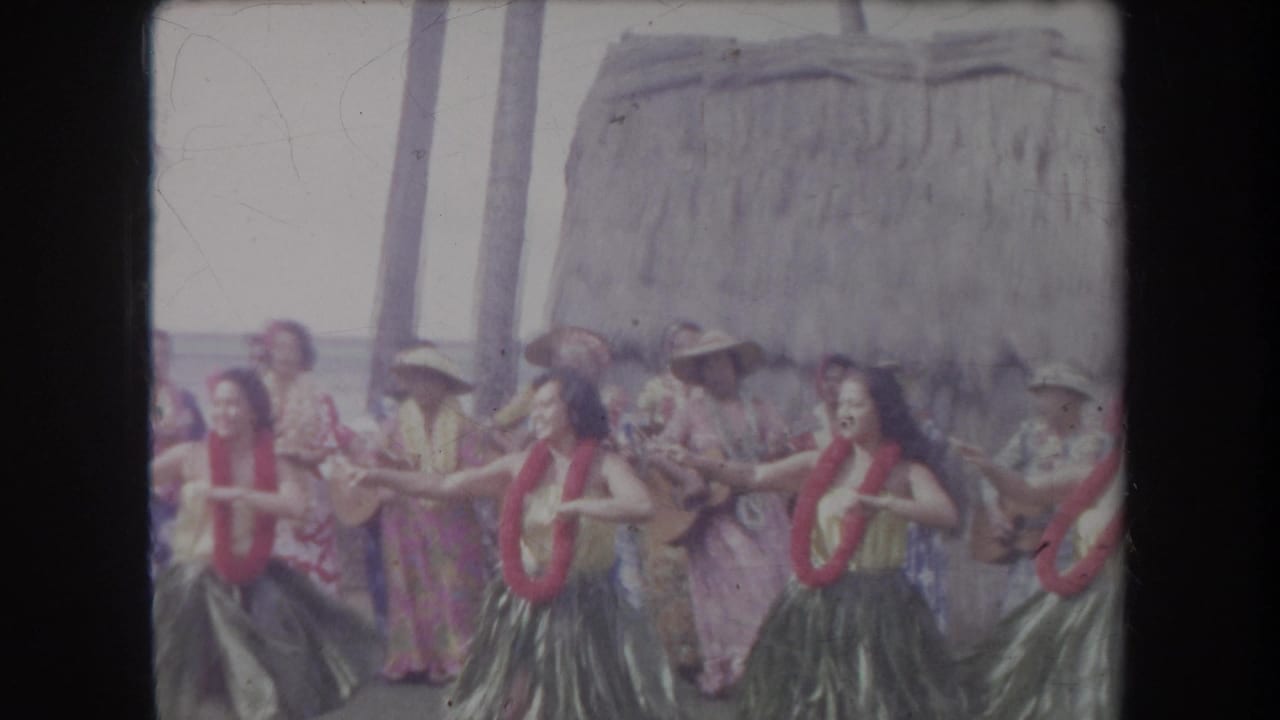 In Hawaii 1969 home-movie footage, hula dancers perform in front of musicians, palm trunks, and a thatched beach structure.