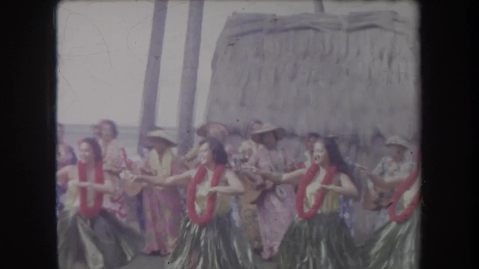 In Hawaii 1969 home-movie footage, hula dancers perform in front of musicians, palm trunks, and a thatched beach structure.