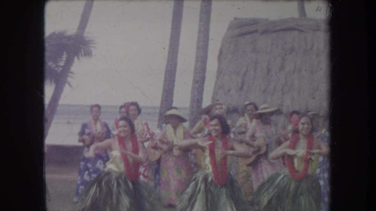 Hula dancers in red leis and grass skirts perform in a Hawaii beach setting with musicians and ocean visible behind them.