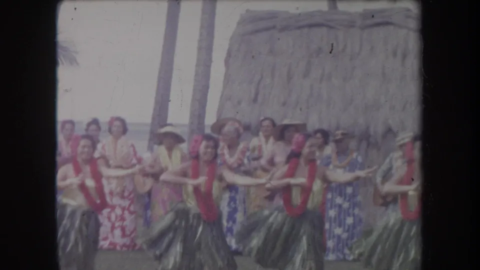 A handheld 8mm frame shows a hula dancer in the foreground with musicians and other performers near a beach hut in Hawaii.