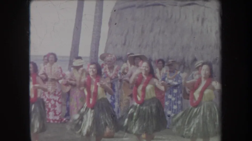 Hula dancers wearing red leis perform in a 1969 Hawaii beach scene, accompanied by musicians in colorful shirts and hats.