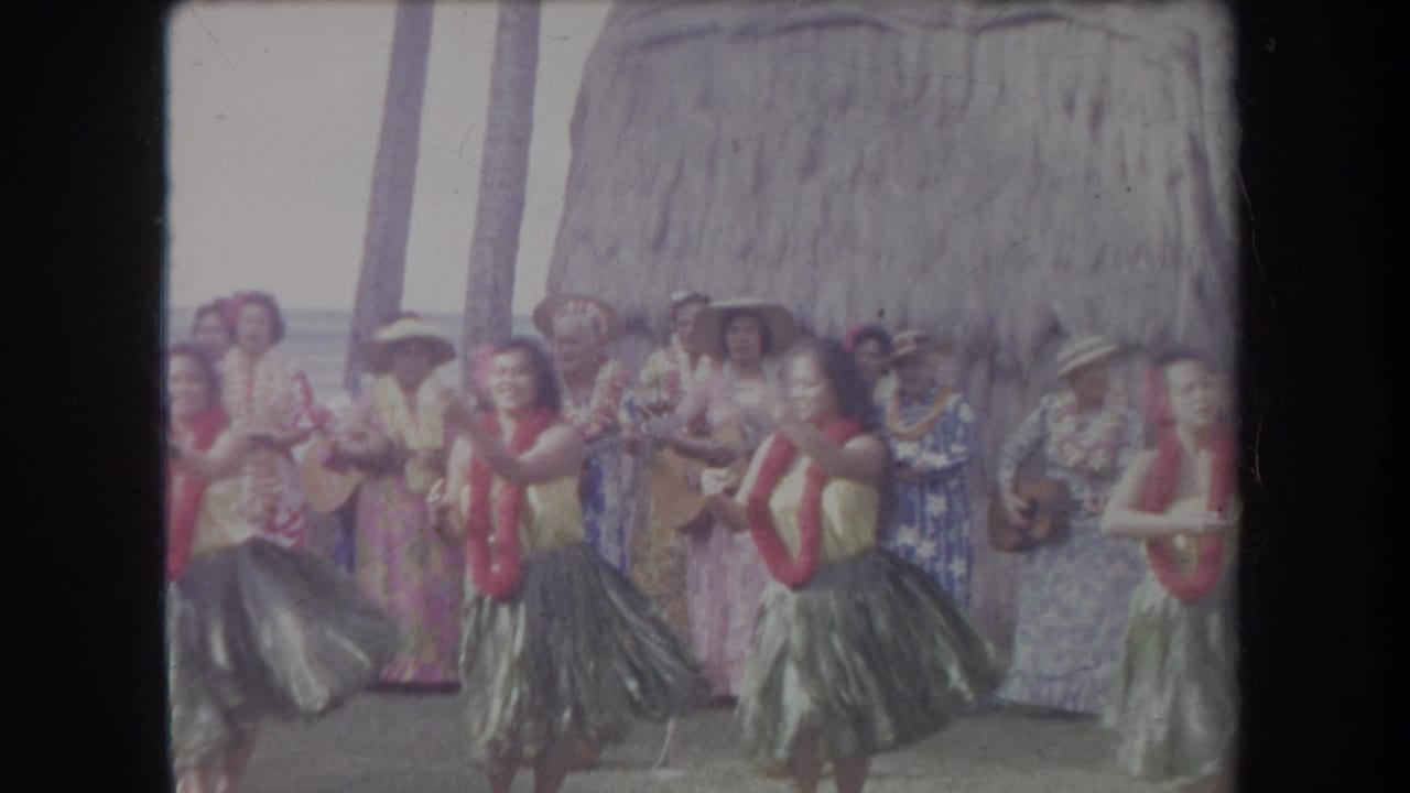1969 Hawaii 8mm frame showing hula dancers with red leis performing in front of ukulele musicians and a thatched backdrop.