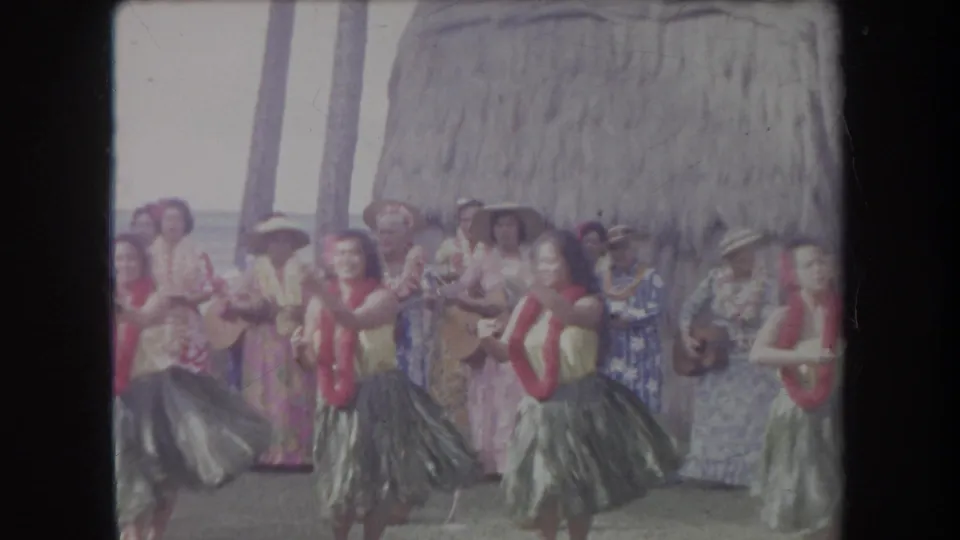 1969 Hawaii 8mm frame showing hula dancers with red leis performing in front of ukulele musicians and a thatched backdrop.