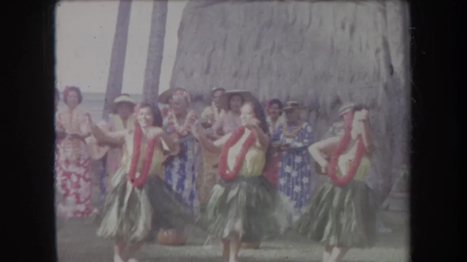 1969 Hawaii 8mm frame with dancers in leis and grass skirts performing before musicians, a gourd, palm trunks, and thatched backdrop.