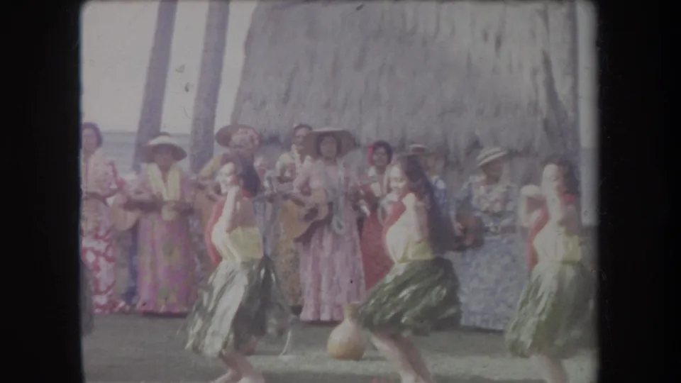 1969 Hawaii 8mm frame showing dancers in motion before a line of musicians with ukuleles and floral dresses near a thatched backdrop.