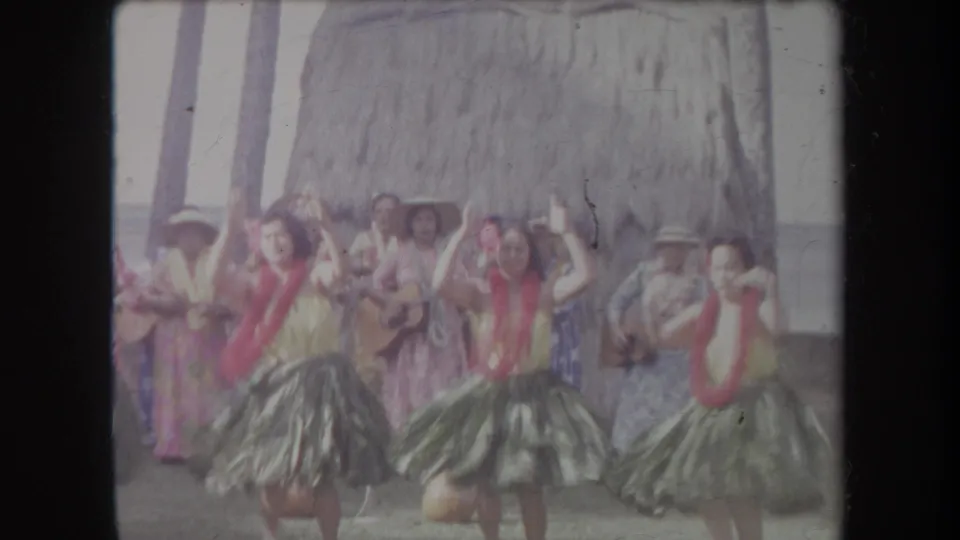 1969 Hawaii 8mm frame of hula dancers raising their arms as musicians play behind them near palm trunks and a thatched backdrop.