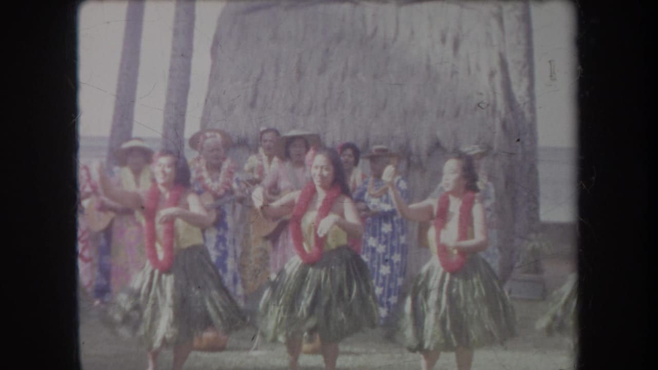 1969 Hawaii 8mm frame showing several women performing hula in grass skirts and red leis before a thatched backdrop.