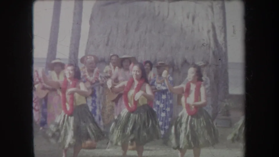 1969 Hawaii 8mm frame showing several women performing hula in grass skirts and red leis before a thatched backdrop.