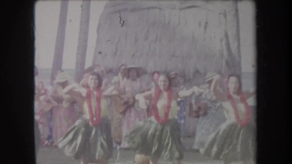 1969 Hawaii 8mm frame showing group hula dancers in motion with musicians or attendants behind them near a thatched hut.
