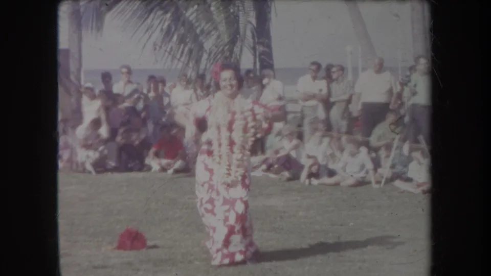 1969 Hawaii home-movie frame of a solo dancer in a floral dress performing on a lawn before a seated beachside audience.
