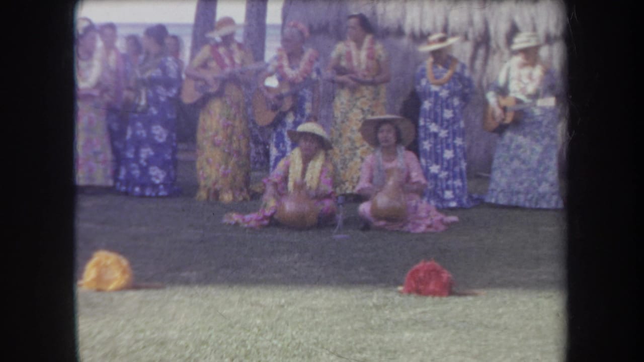 1969 Hawaii 8mm frame of women in leis and floral muumuus performing music outdoors with seated performers in front.