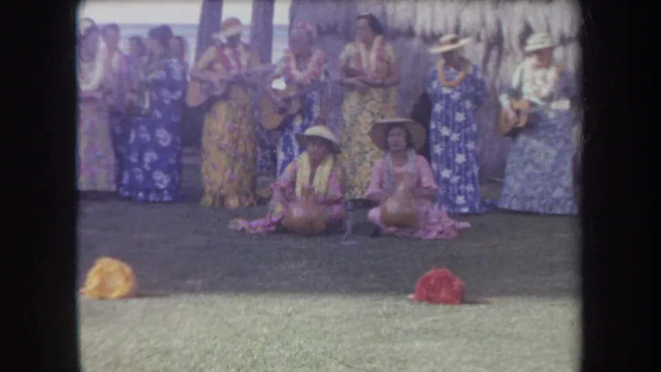 1969 Hawaii 8mm frame of women in leis and floral muumuus performing music outdoors with seated performers in front.