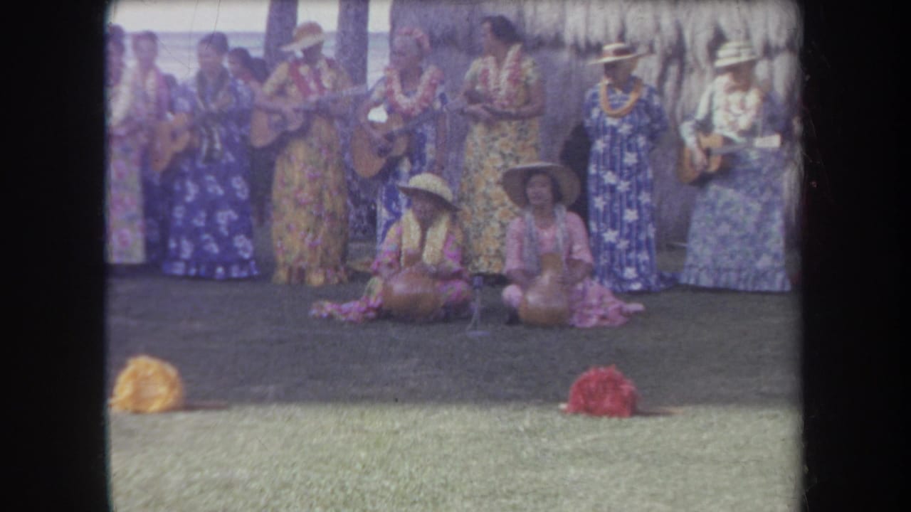 Musicians and seated performers remain grouped on the outdoor stage near a thatched backdrop.