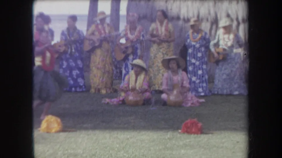 1969 Hawaii 8mm frame with a young hula dancer entering the foreground while women play music behind her.