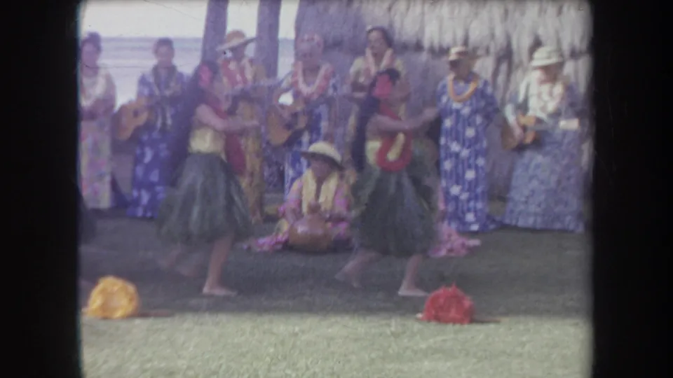 1969 Hawaii home-movie frame of two young hula dancers in grass skirts moving before women with ukuleles and leis.