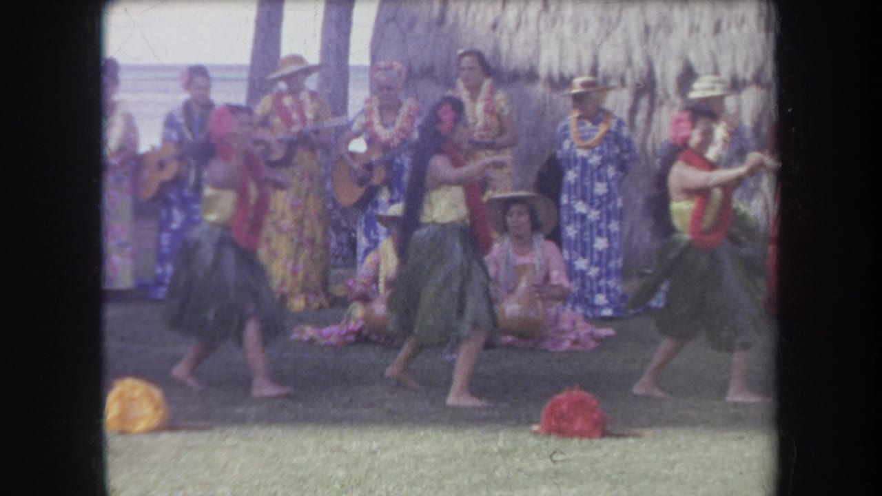 Hula dancers perform on grass in front of musicians and a thatched structure.