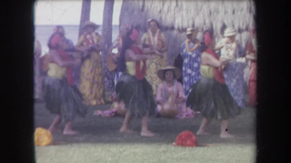 The 8mm frame shows hula dancers mid-motion with red leis, while ukulele players and other performers stand near a thatched wall.