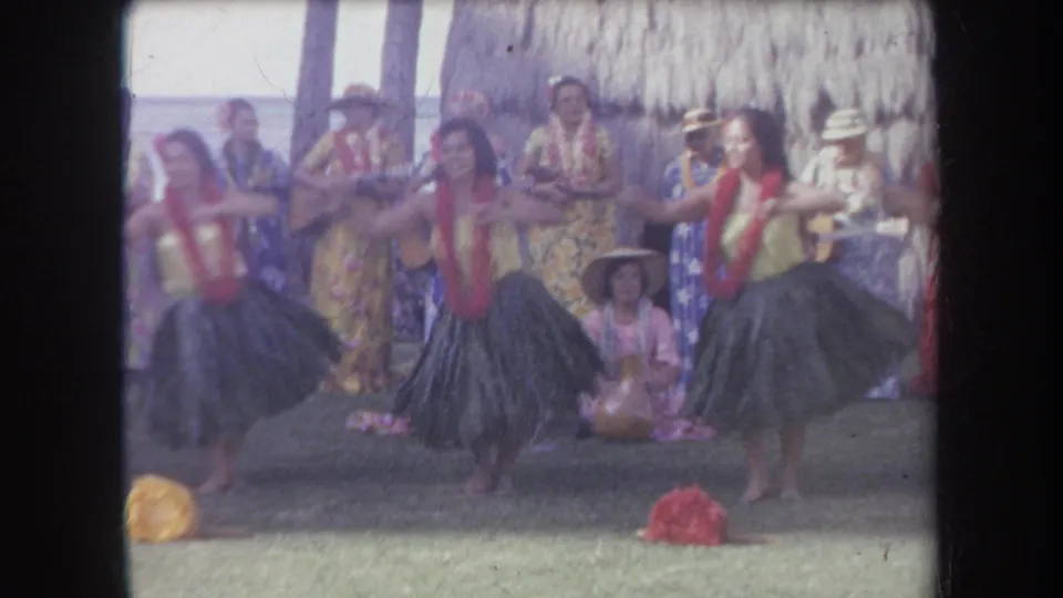 A hula dancer faces forward during the 1969 Hawaii performance, with other dancers, musicians, leis, and a gourd instrument visible behind.
