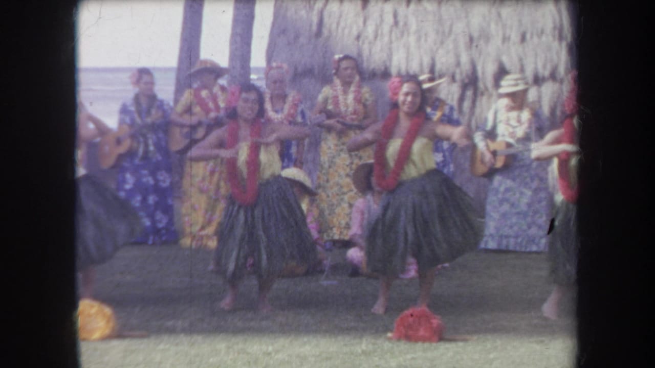 1969 Hawaii home-movie frame showing hula dancers in grass skirts and red leis with musicians behind them near a thatched shelter.