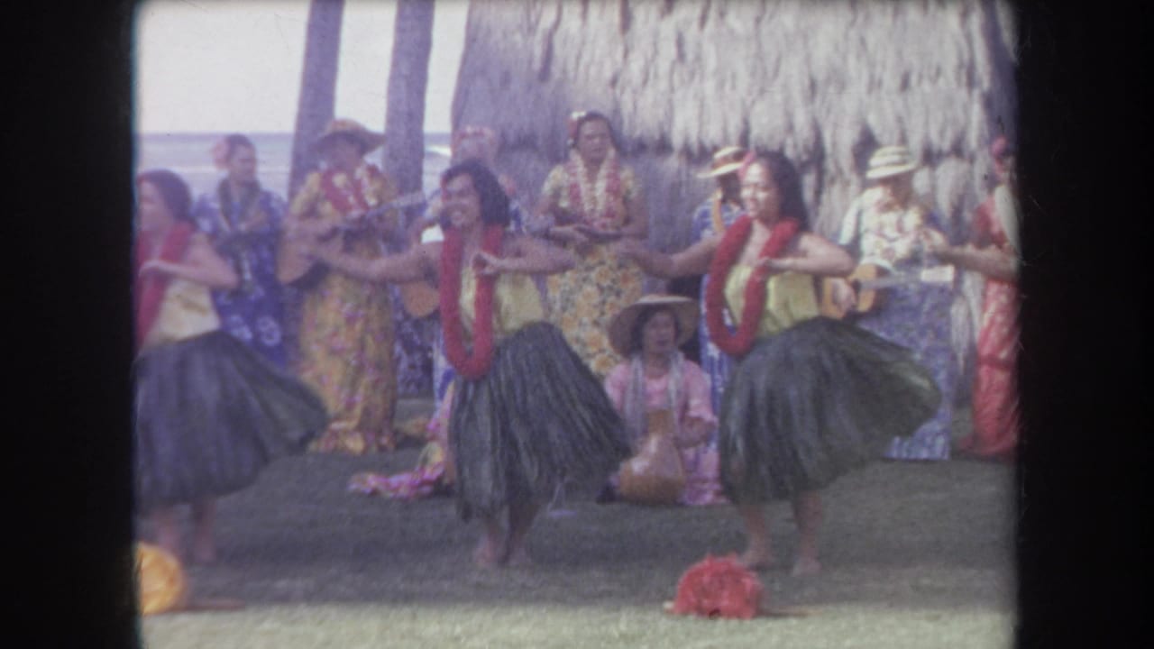 Hula dancers extend their arms while musicians and seated spectators are behind them.