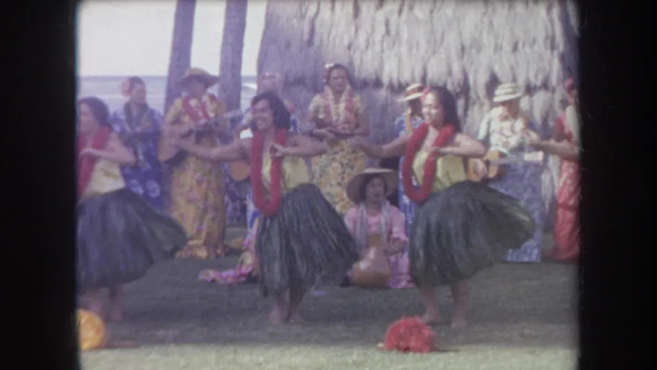 1969 Hawaii frame with hula dancers mid-motion, guitar players, floral-costumed performers, and a seated spectator near the stage area.