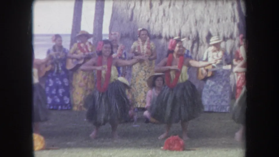 1969 Hawaii home-movie frame of two hula dancers performing before musicians in floral clothing at a beachside thatched shelter.