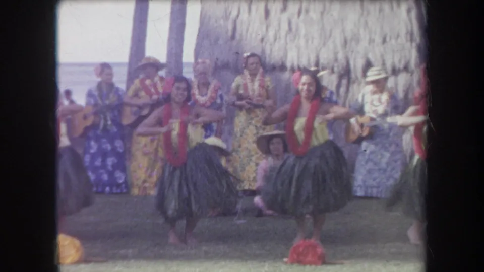 1969 Hawaii frame showing a smiling hula dancer, another dancer beside her, musicians, a seated child-like spectator, and beach scenery.