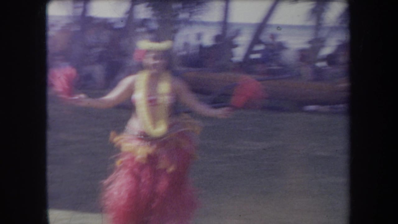 1969 Hawaii 8mm frame of a hula dancer in lei and grass skirt, blurred in motion before a shaded beachside audience area.