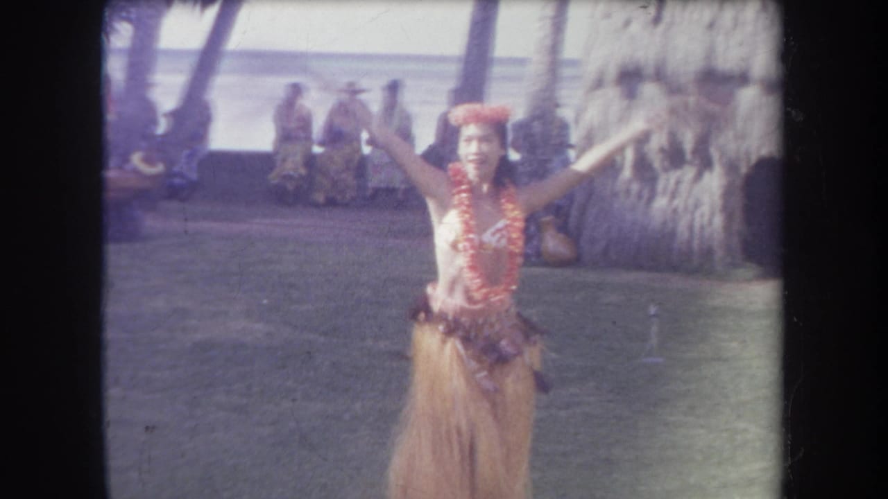 1969 Hawaii home-movie frame showing a hula dancer in floral adornments and grass skirt performing beside a thatched structure with onlookers behind her.