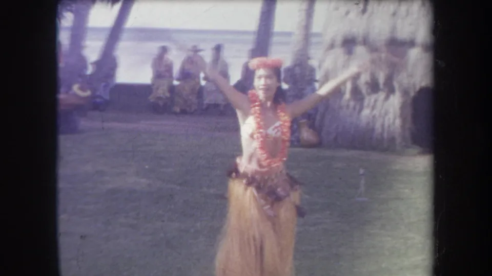 1969 Hawaii home-movie frame showing a hula dancer in floral adornments and grass skirt performing beside a thatched structure with onlookers behind her.
