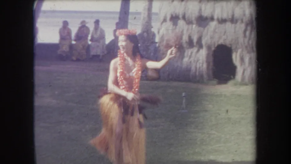 1969 Hawaii 8mm frame of a hula dancer gesturing beside a thatched wall, with several audience members seated near palm trees in the background.