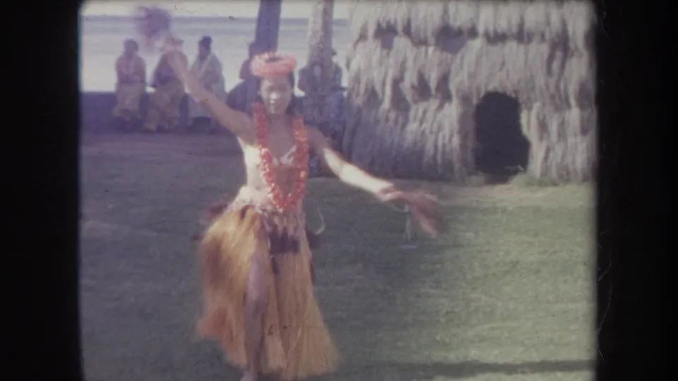 1969 Hawaii home-movie frame showing a hula dancer in a lei and grass skirt extending an arm near a thatched hut as visitors sit in the shade.