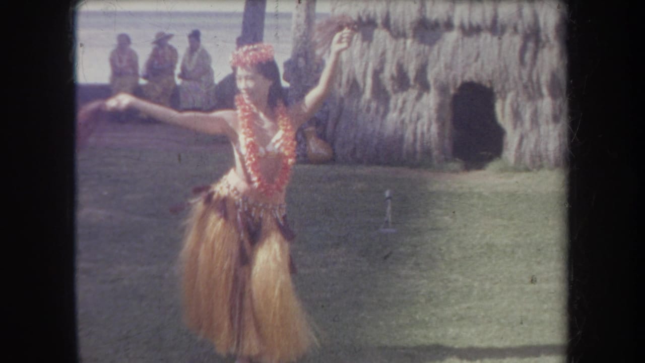 1969 Hawaii 8mm frame showing a hula dancer facing camera in a grass skirt, red lei, and floral headpiece beside a thatched hut.