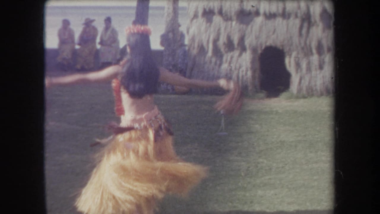 Hula dancer seen from behind with arms extended while spinning on grass.