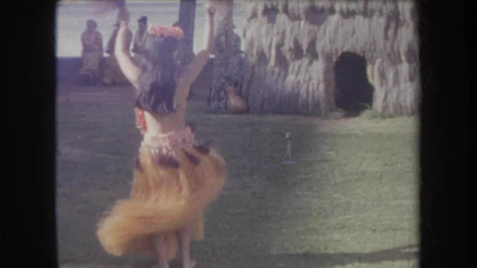 1969 Hawaii 8mm frame showing the hula dancer from behind with raised arms, grass skirt in motion, seated onlookers, and a thatched structure.