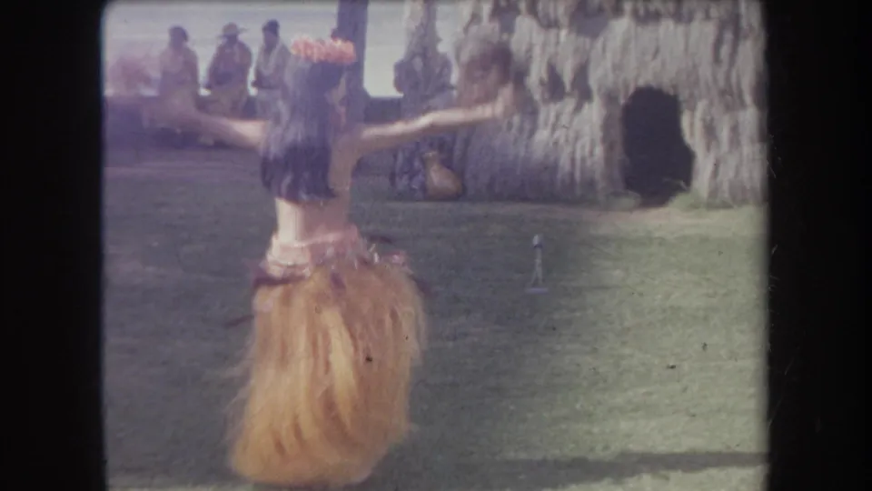 1969 Hawaii 8mm frame showing the dancer from behind with arms spread, wearing a grass skirt and lei near a thatched hut and lawn audience.