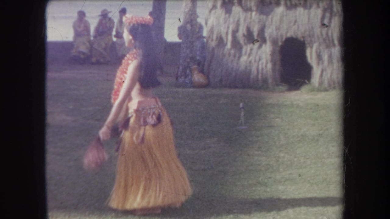 1969 Hawaii 8mm frame of a hula dancer in grass skirt and flower lei performing outdoors near seated spectators, palms, and a thatched wall.