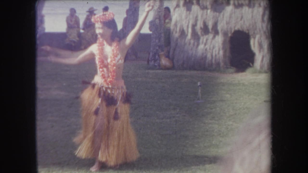 Hula dancer raises one arm while performing on grass, with seated spectators and a thatched structure behind her.
